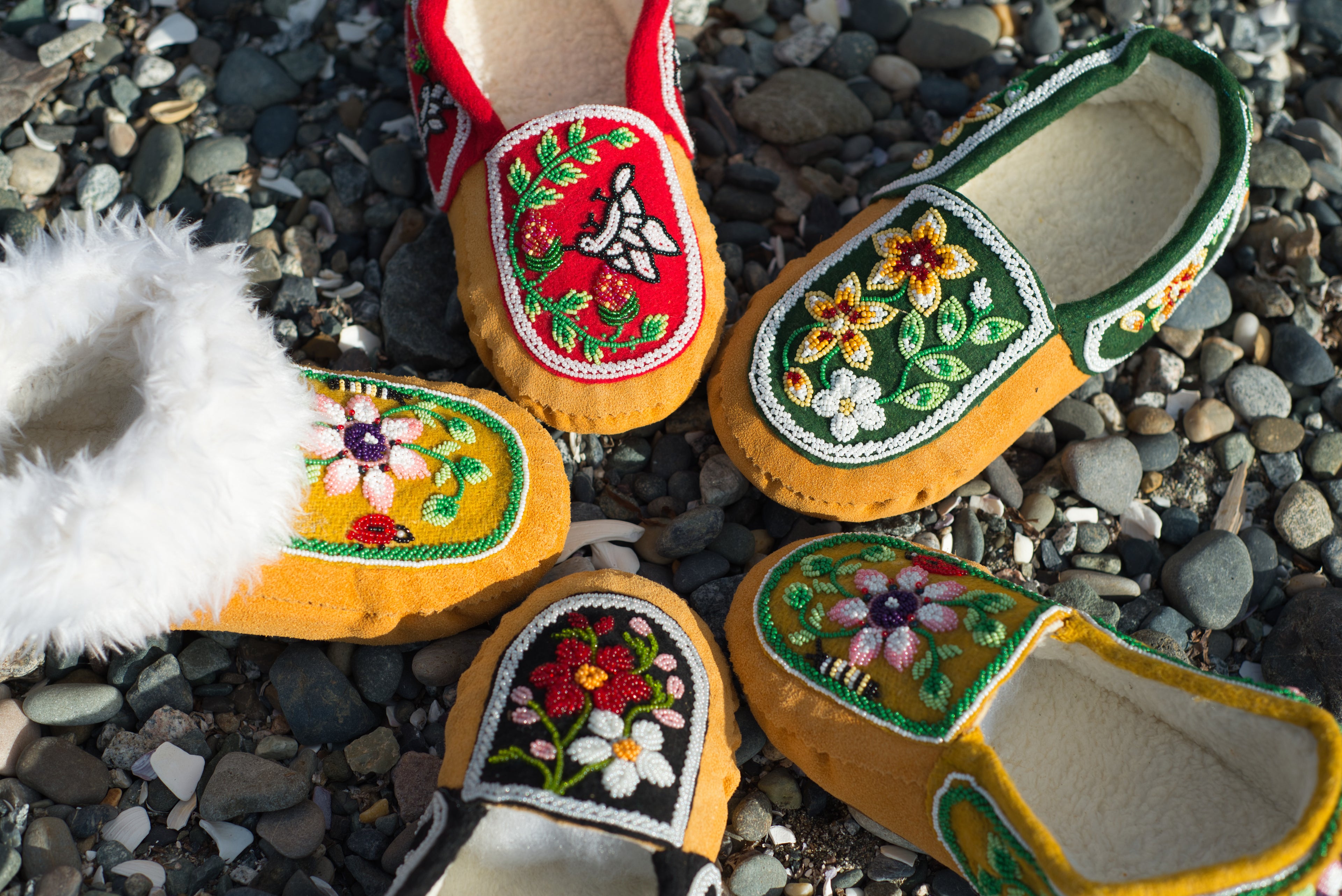 A collection of five different handmade beaded moccasins arranged in a circle on a rocky beach.