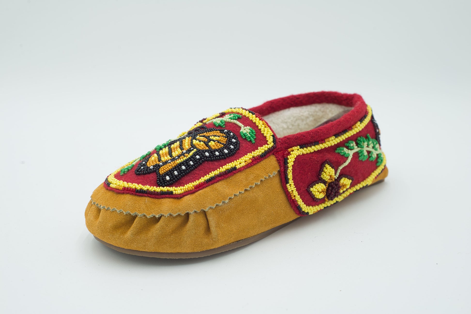 A single tan cow suede slip-on moccasin with a raised 3D beaded butterfly in gold, black, and green on a crimson vamp, fleece-lined, shown on a white background.