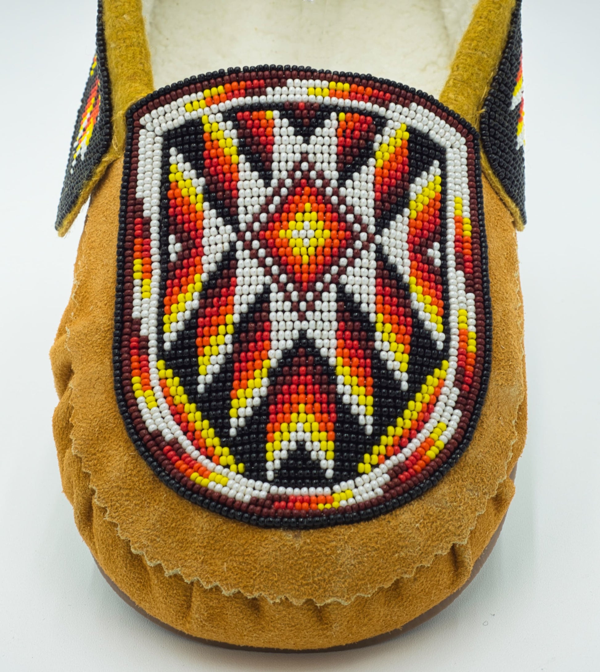 A single tan cow suede slip-on moccasin with red, orange, and yellow thunderbird flame beadwork, a mustard-gold fleece-lined collar, and rubber sole, shown on a white background. zoomed in. deatailed work