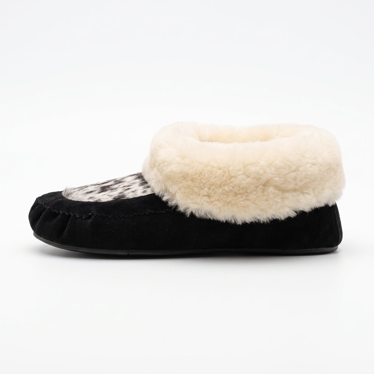 Side view of a black cow suede moccasin bootie with a speckled white and black hair-on cowhide vamp and a thick cream real sheepskin fold-over cuff, on a white background.