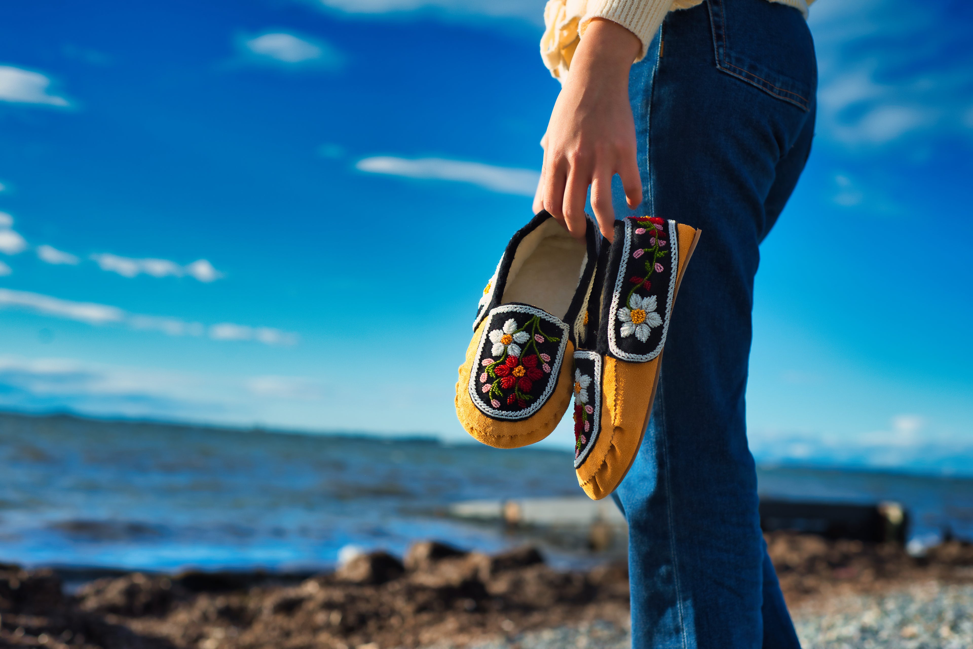 A person holding handmade floral beaded moccasins on a sunny Canadian beach.